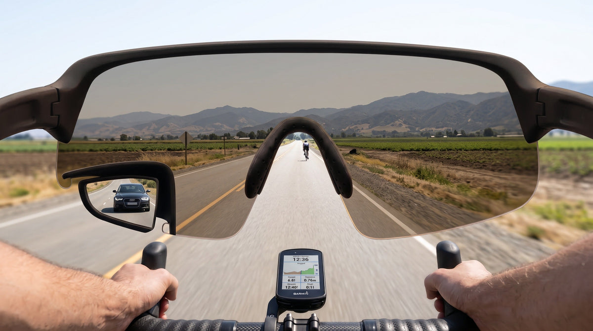 Point-of-view through TriEye The View on a country road: one shield lens with a small rear-view mirror set into its lower left corner, a car visible in the mirror, handlebars and bike computer below