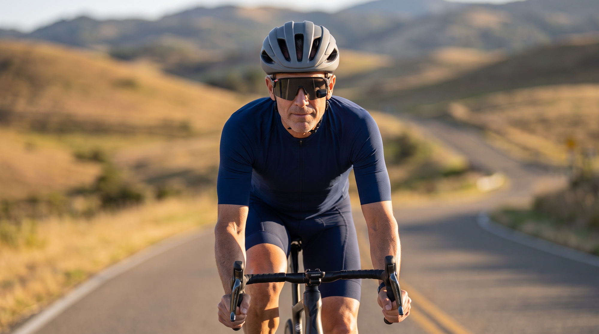 Road cyclist in a plain helmet and sport wraparound sunglasses with an in-lens rear-view mirror, riding on an open sunlit road
