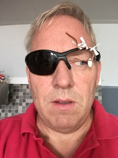 Een man met een rood overhemd en een donkere zonnebril met kapot glas, bijeengehouden door een witte stropdas, staat binnen voor een betegelde muur.