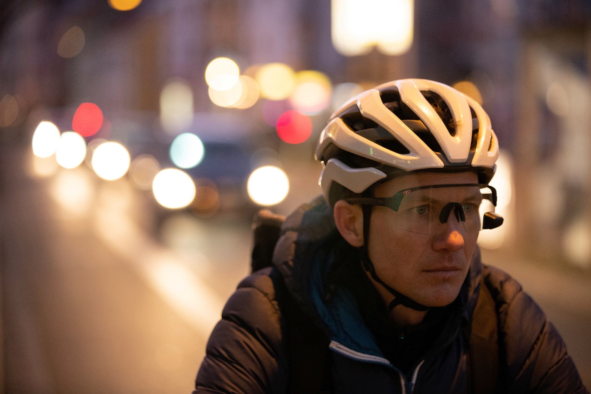 Een persoon met een witte helm, bril en jas fietst in een stad in de schemering, met vage koplampen van auto's en straatverlichting op de achtergrond.