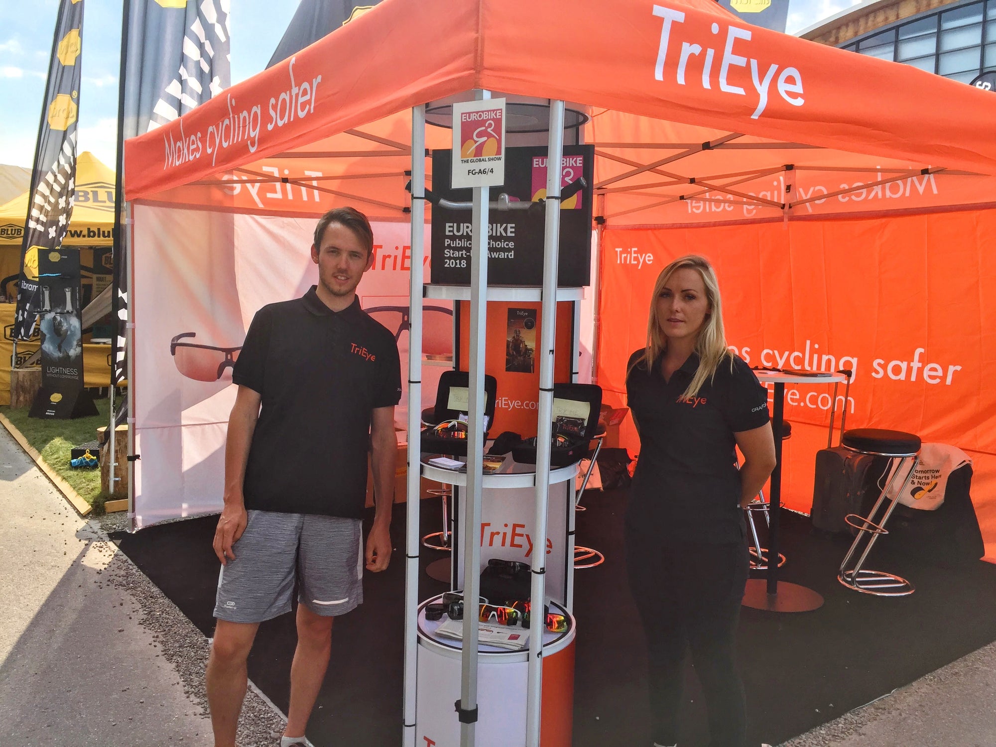 Twee mensen staan onder een oranje tent met TriEye-logo op een buitenevenement en tonen fietsproducten op een stand. Op de achtergrond zijn spandoeken, stoelen en de omgeving van het evenement te zien.