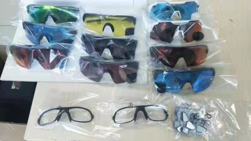 Tien paar sportzonnebrillen met verschillende gekleurde glazen staan in drie rijen, elk paar in een plastic zakje. Onderaan liggen twee doorzichtige monturen en een zakje met neuspads of accessoires.