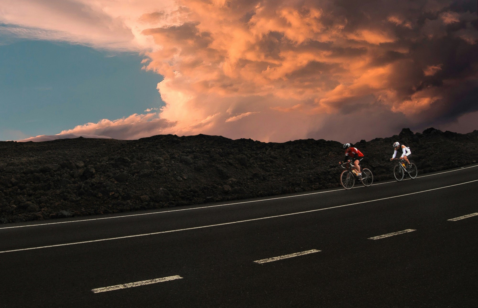 Twee fietsers rijden op een lege weg langs donker rotsachtig terrein onder een dramatische lucht vol oranje en roze wolken bij zonsondergang.